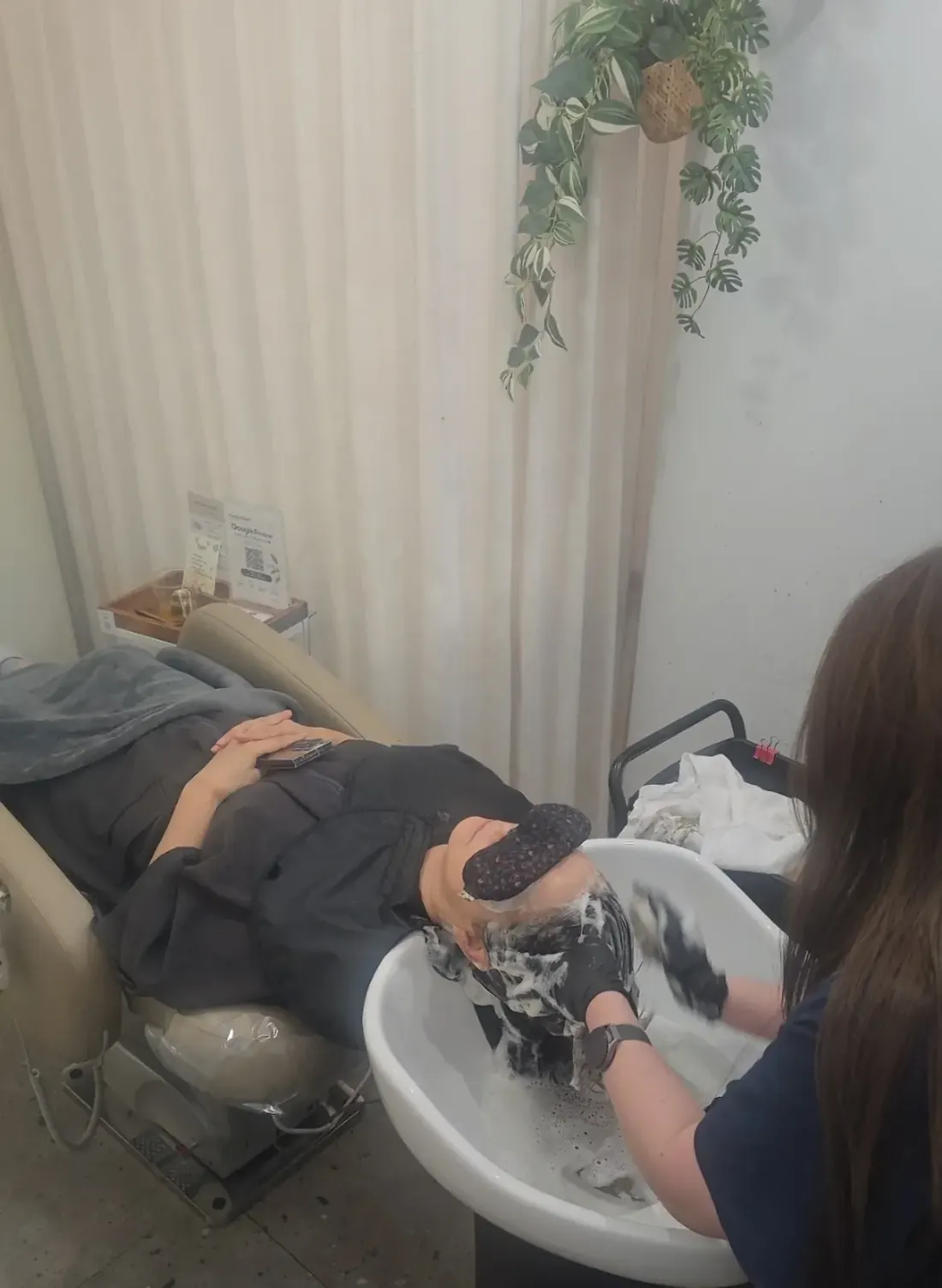 샴푸대에 누워 눈가림 패드를 하고 두피 관리를 받는 고객과 관리 담당자